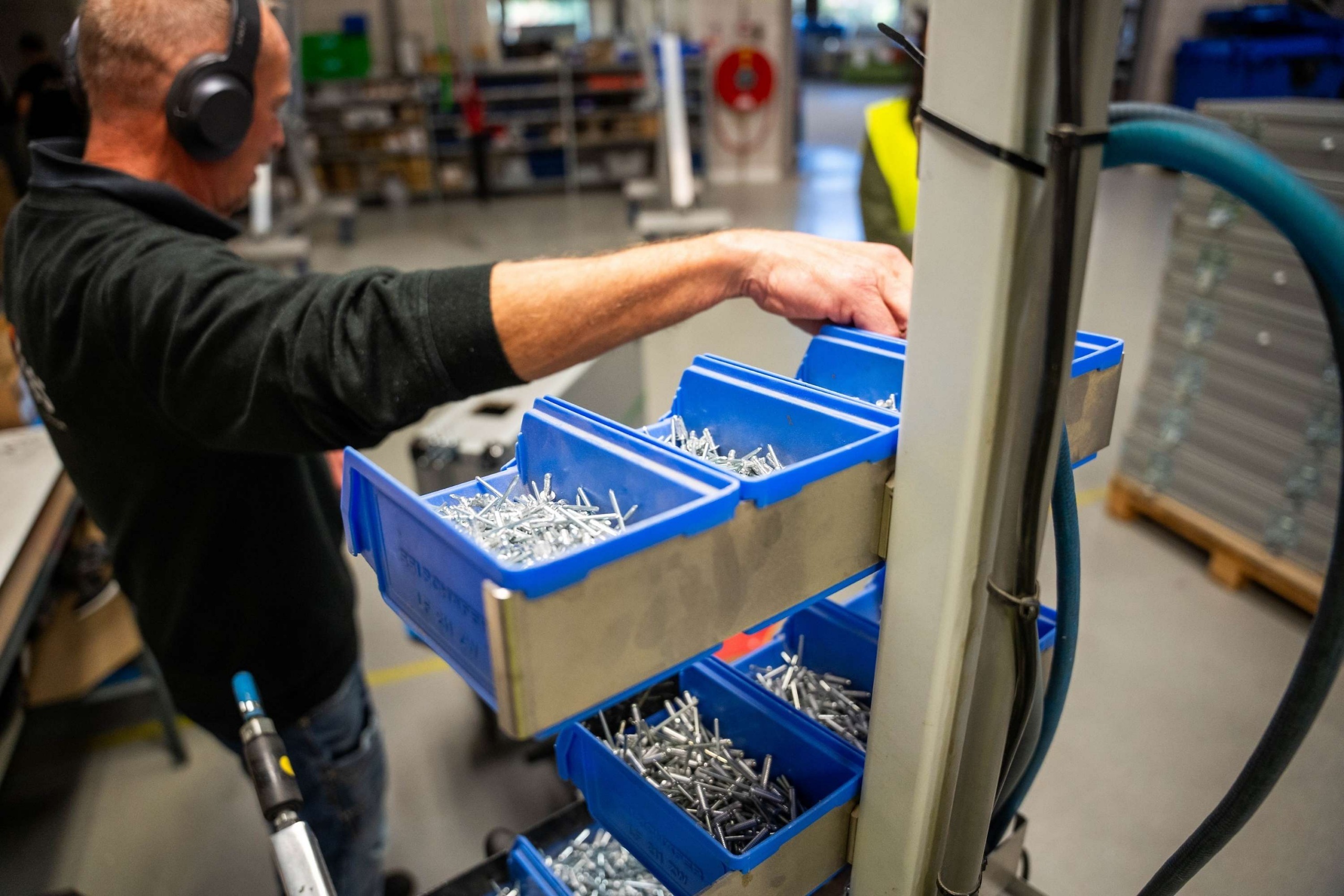 Medewerker van Faes pakt metalen bevestigingsmateriaal uit blauwe onderdelenbakken in de productiehal, passend bij de montage van robuuste scharniermechanismen.