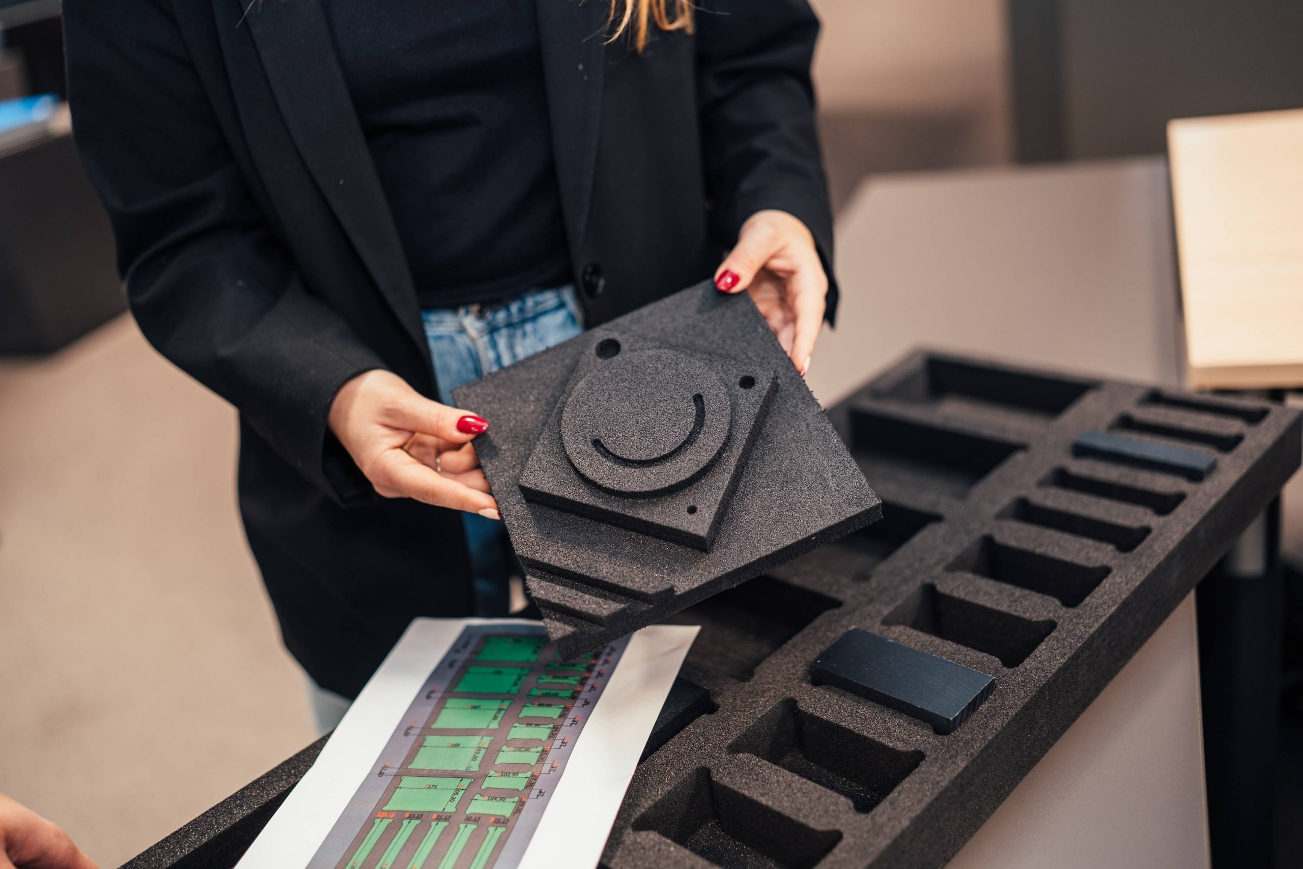 Medewerker van Faes houdt een zwarte foam inzet met uitgesneden vormen boven een maatwerk foam interieur met compartimenten, passend bij het ontwerpen van beschermende interieurs voor kwetsbare onderdelen.