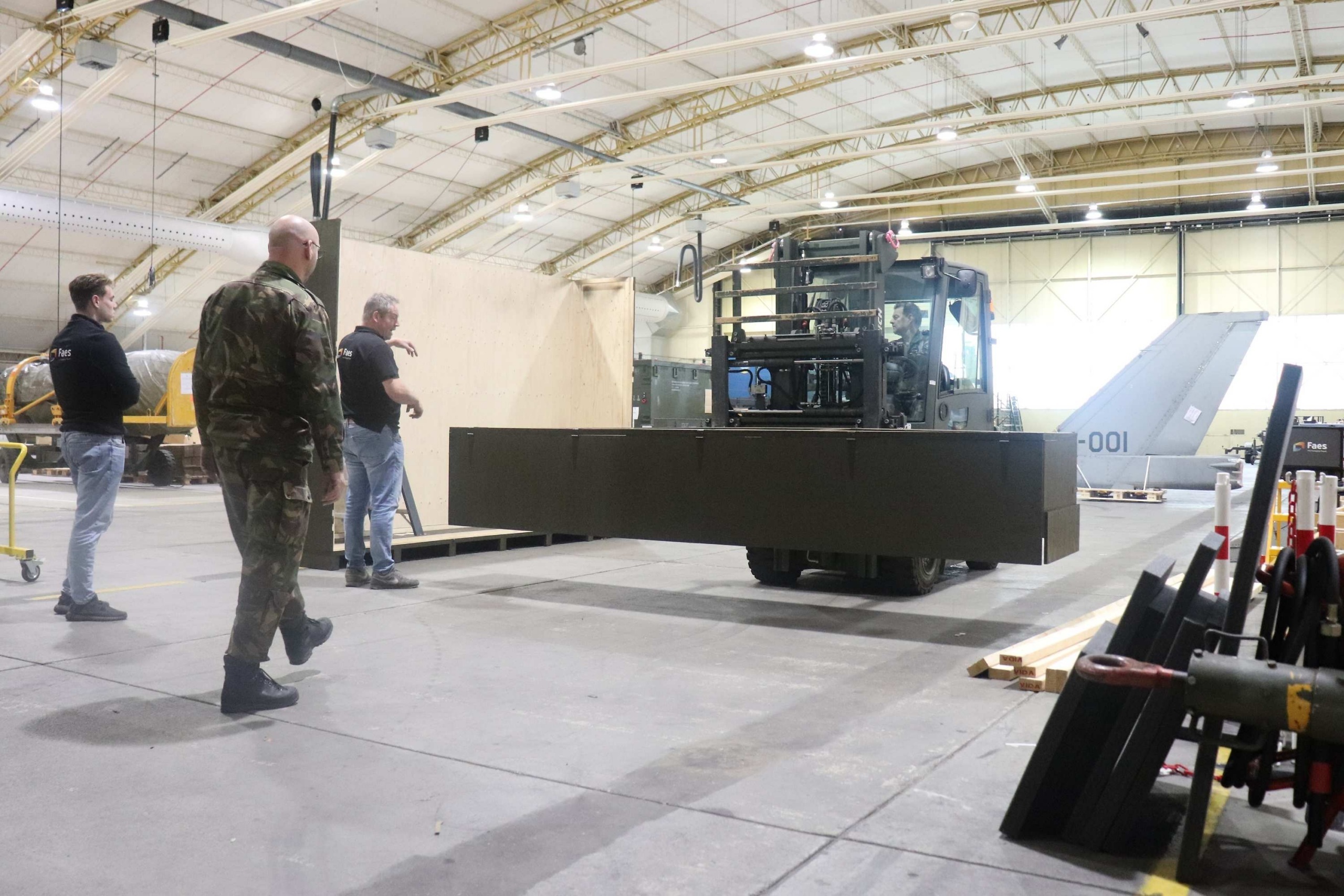 Medewerkers van Faes en een militair begeleiden in een hangar een grote transportverpakking op een heftruck, passend bij veilig gebruik en onderhoud onder veeleisende omstandigheden.