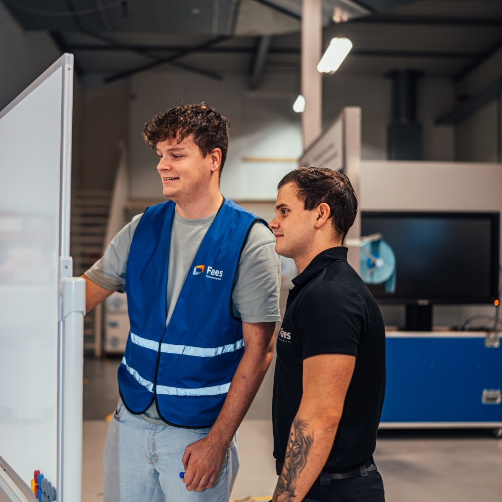 Twee medewerkers van Faes bespreken ideeën bij een whiteboard in de productieomgeving, passend bij efficiënte verpakkingslogistiek en emissiereductie binnen FuelEU Maritime.