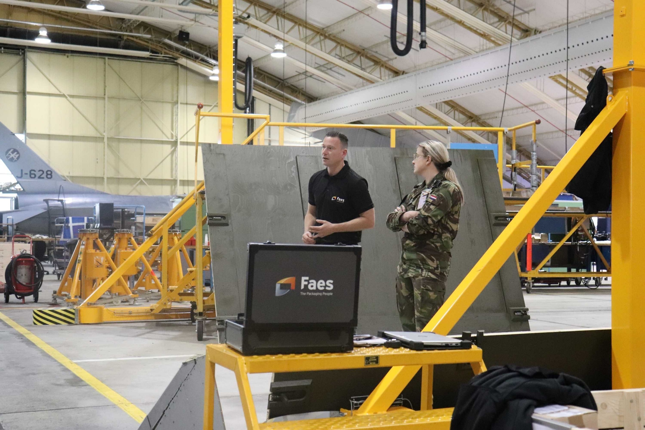 Medewerker van Faes bespreekt in een militaire onderhoudshal een beschermende transportverpakking met een defensiemedewerker, met een vliegtuig op de achtergrond.