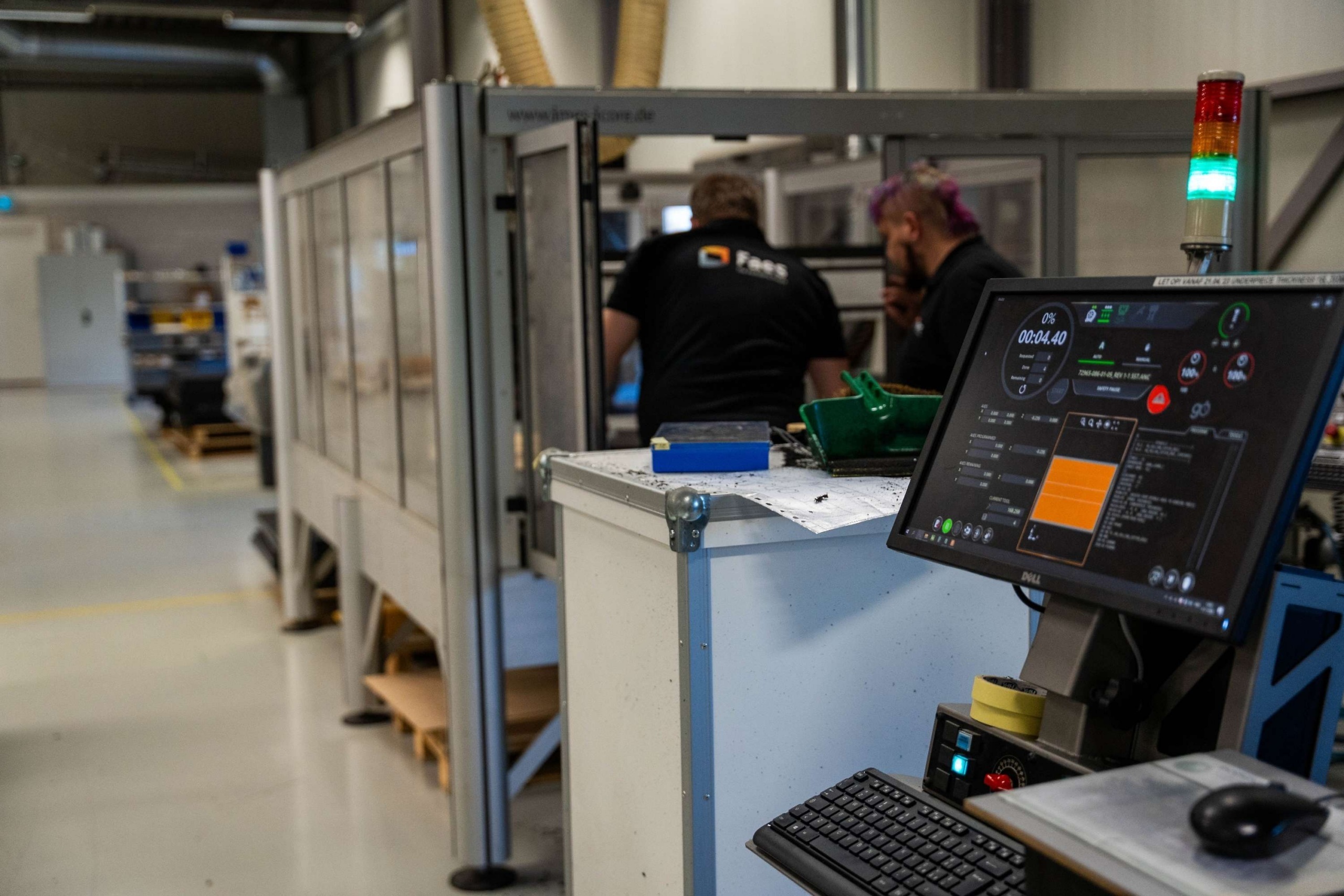 In de productiehal van Faes staat een bedieningsscherm naast een afgeschermde productiemachine, terwijl twee medewerkers op de achtergrond aan het werk zijn.