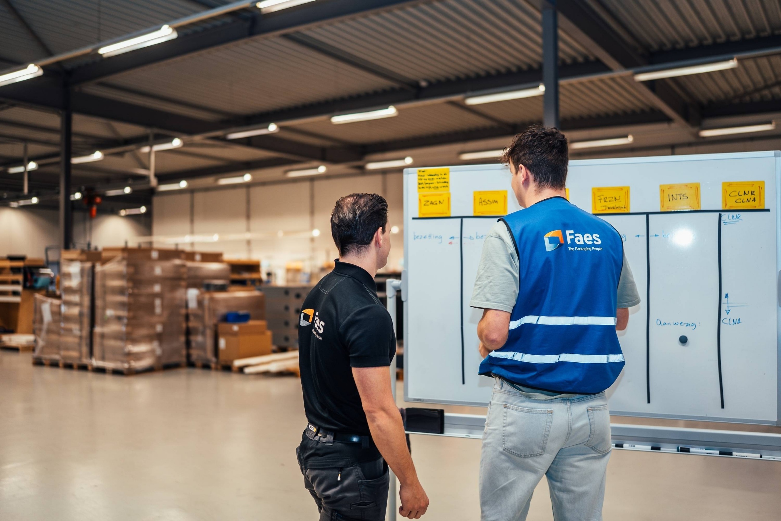 Twee medewerkers van Faes bespreken in de productiehal een planning op een whiteboard, als onderdeel van een gestructureerde aanpak om Dead-on-Arrival risico’s in de supply chain te voorkomen.