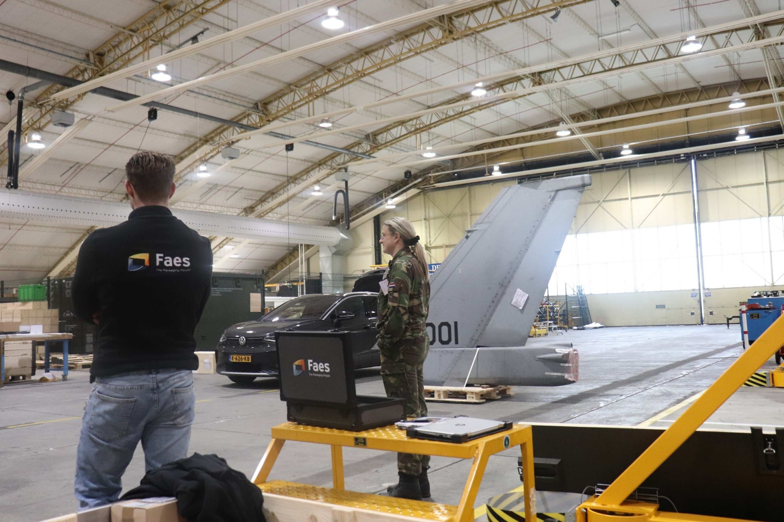 Medewerker van Faes staat met een defensiemedewerker in een vliegtuighangar bij een robuuste transportkist, passend bij het gebruik van beschermende kisten voor militaire uitrusting en munitie.