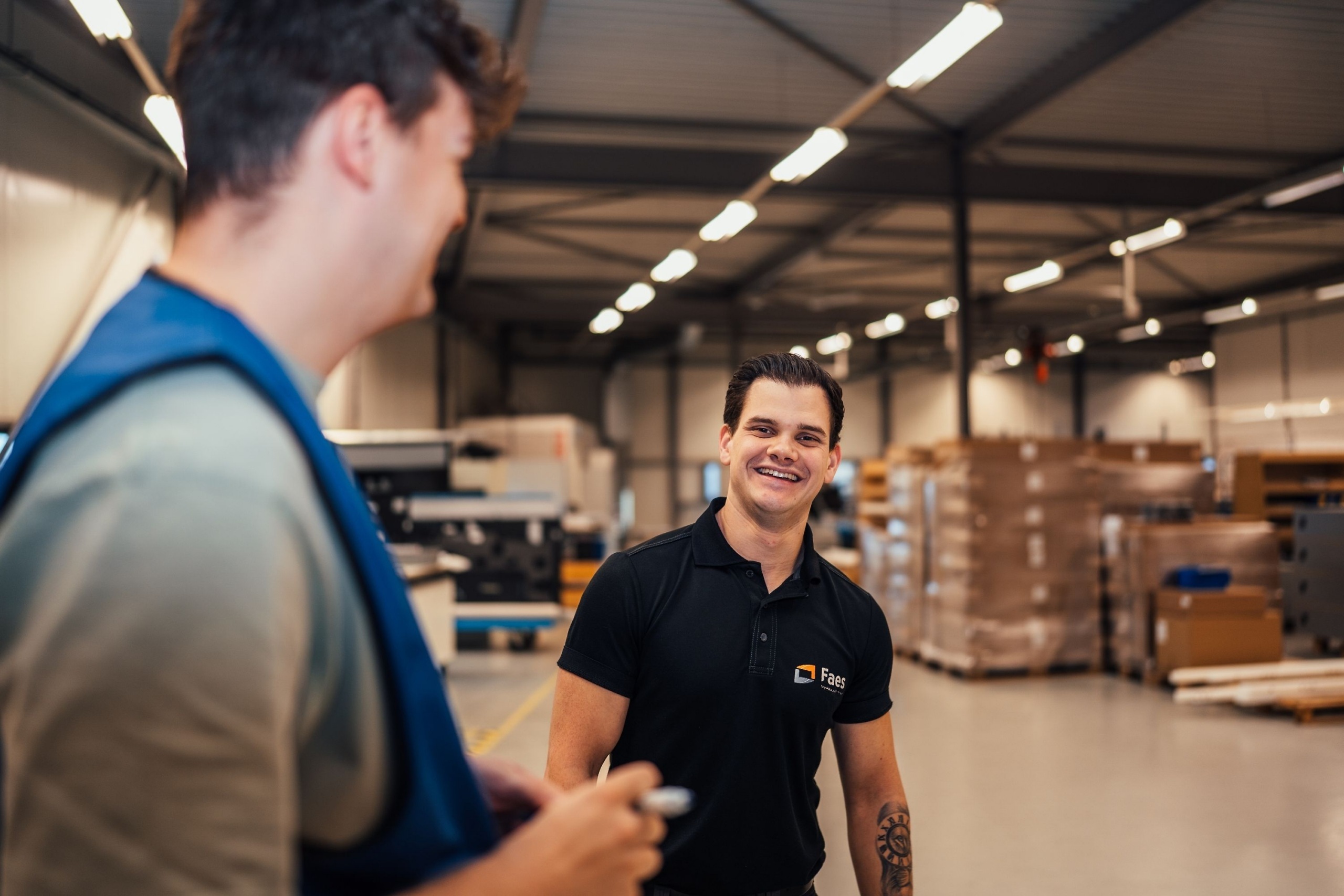 Medewerkers van Faes overleggen in de productiehal tussen pallets en verpakkingsmaterialen, passend bij het maken van kostenbewuste verpakkingskeuzes op basis van TCO.